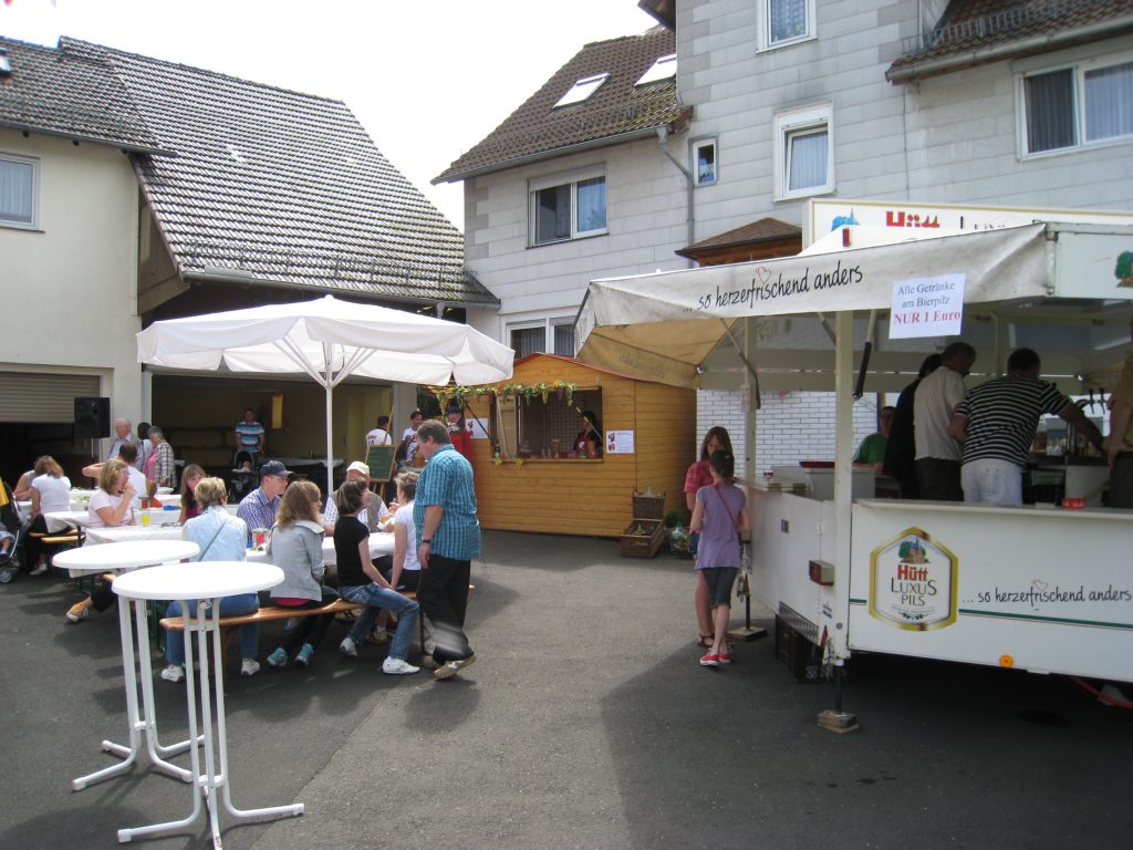 Hof vor Holzbach 16 mit feiernden Menschen, Bierzeltgarnituren, Stehstichen, Bierpilz und weiterem Getränkestand